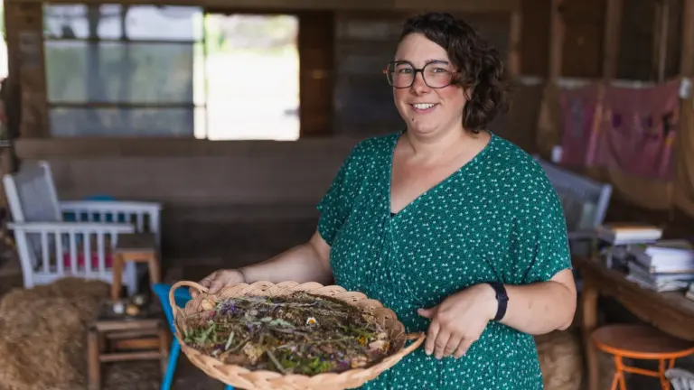Une femme souriante aux cheveux courts tenant un plateau en osier rempli de fleurs séchées.