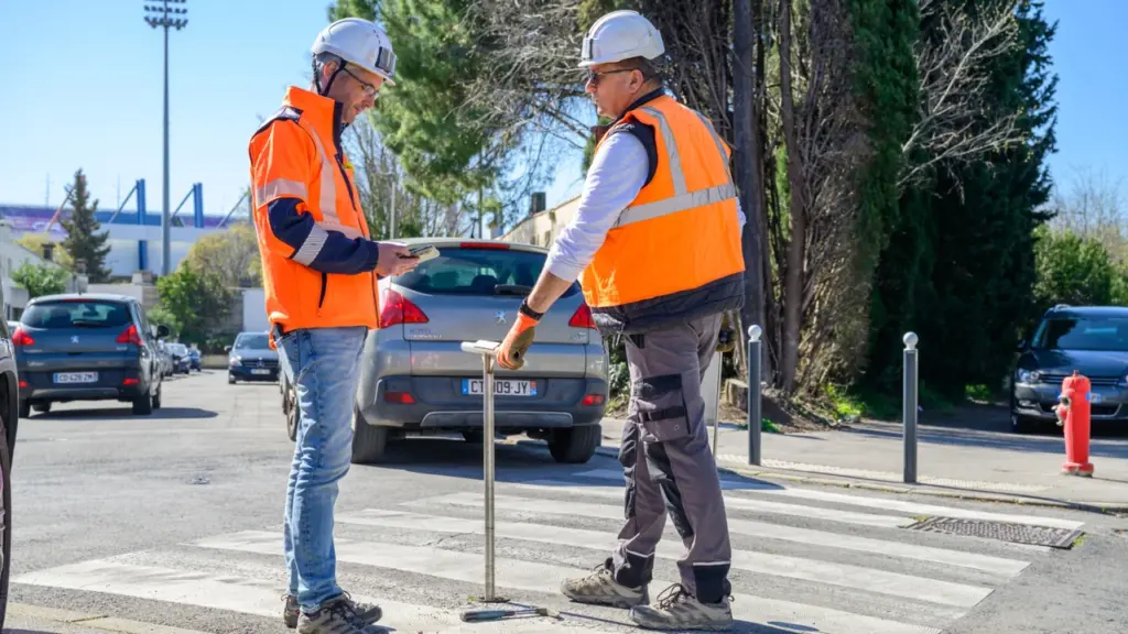 Deux ouvriers en gilets orange et casques de chantier inspectent un passage piéton urbain.