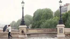Une femme avec un parapluie noir marche sur un pont parisien par temps pluvieux.