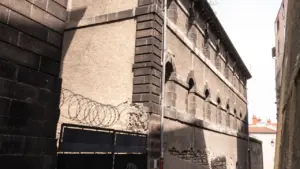 Une rue étroite longe un bâtiment ancien en pierre avec des barbelés sur le toit.